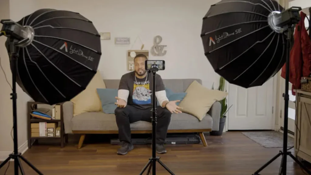 Дві круглі софтбокси Aputure Light Dome SE на стійках освітлюють чоловіка на сірому дивані у домашній вітальні, смартфон на штативі знімає інтерв'ю