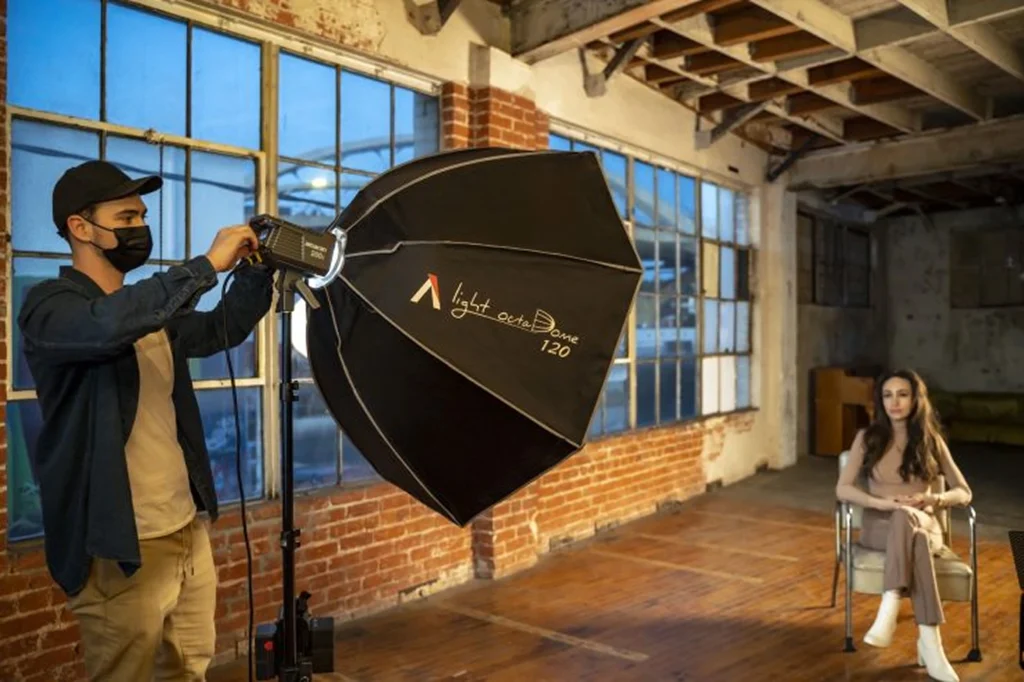 Восьмикутний софтбокс Aputure Light octaDome 120 на студійному прожекторі, оператор у масці спрямовує велике м'яке світло на модель у лофті з цегляними стінами і великими вікнами